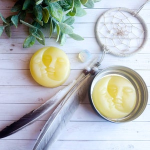 May include: Two yellow wax melts shaped like a crescent moon with a face on each. The melts are on a white wooden surface with a dream catcher and feathers.