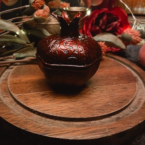Puede incluir: Una peque&ntilde;a caja de madera de granada roja, tallada de forma intrincada, se encuentra sobre una superficie de madera. La caja tiene una tapa y est&aacute; decorada con un patr&oacute;n floral.