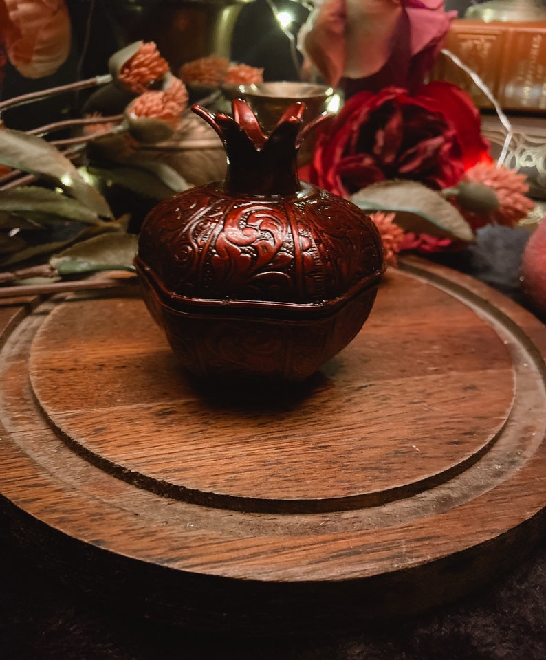 Puede incluir: Una peque&ntilde;a caja de madera de granada roja, tallada de forma intrincada, se encuentra sobre una superficie de madera. La caja tiene una tapa y est&aacute; decorada con un patr&oacute;n floral.