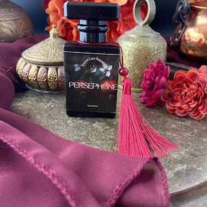 K&ouml;nnte beinhalten: Eine schwarze Glasflasche mit Parf&uuml;m und einem roten Quasten. Die Flasche hat ein Etikett mit der Aufschrift "Secret Lore Botanicals" und "Persephone".