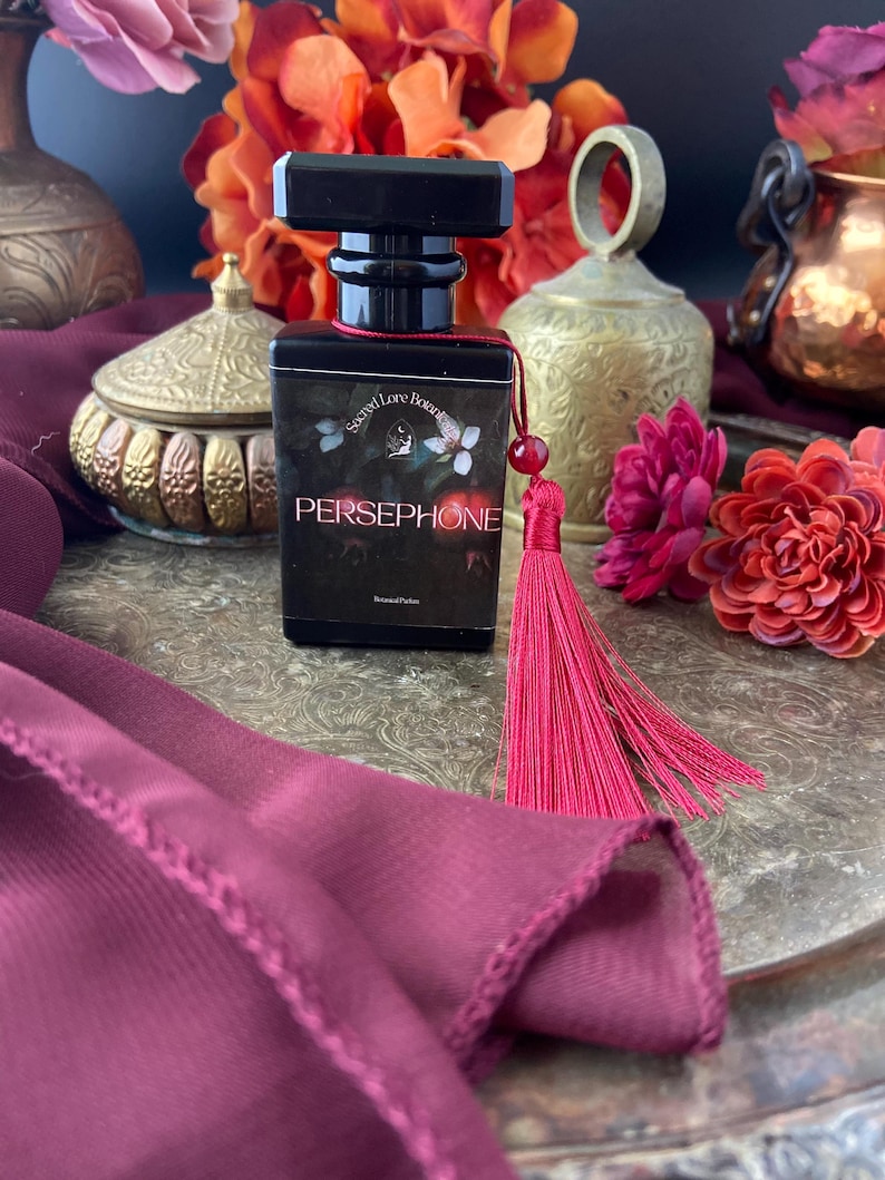 K&ouml;nnte beinhalten: Eine schwarze Glasflasche mit Parf&uuml;m und einem roten Quasten. Die Flasche hat ein Etikett mit der Aufschrift "Secret Lore Botanicals" und "Persephone".
