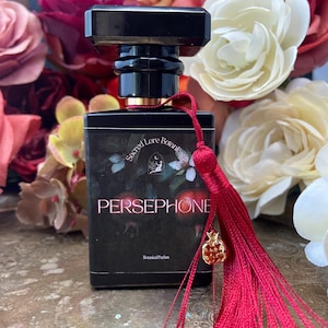 K&ouml;nnte beinhalten: Eine schwarze Glasflasche mit Parf&uuml;m, einem roten Quasten und einem goldenen Granatapfel-Anh&auml;nger. Die Flasche ist mit "Sacred Lore Botanicals Persephone Botanical Parfum" beschriftet.