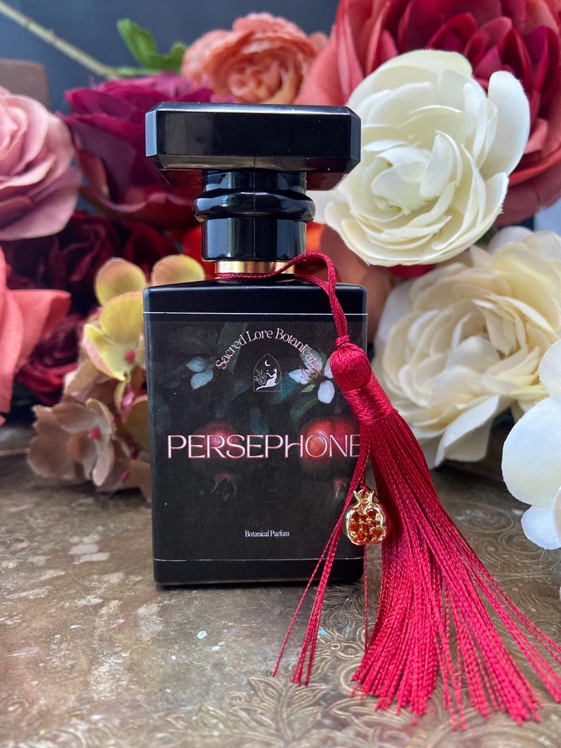 K&ouml;nnte beinhalten: Eine schwarze Glasflasche mit Parf&uuml;m, einem roten Quasten und einem goldenen Granatapfel-Anh&auml;nger. Die Flasche ist mit "Sacred Lore Botanicals Persephone Botanical Parfum" beschriftet.