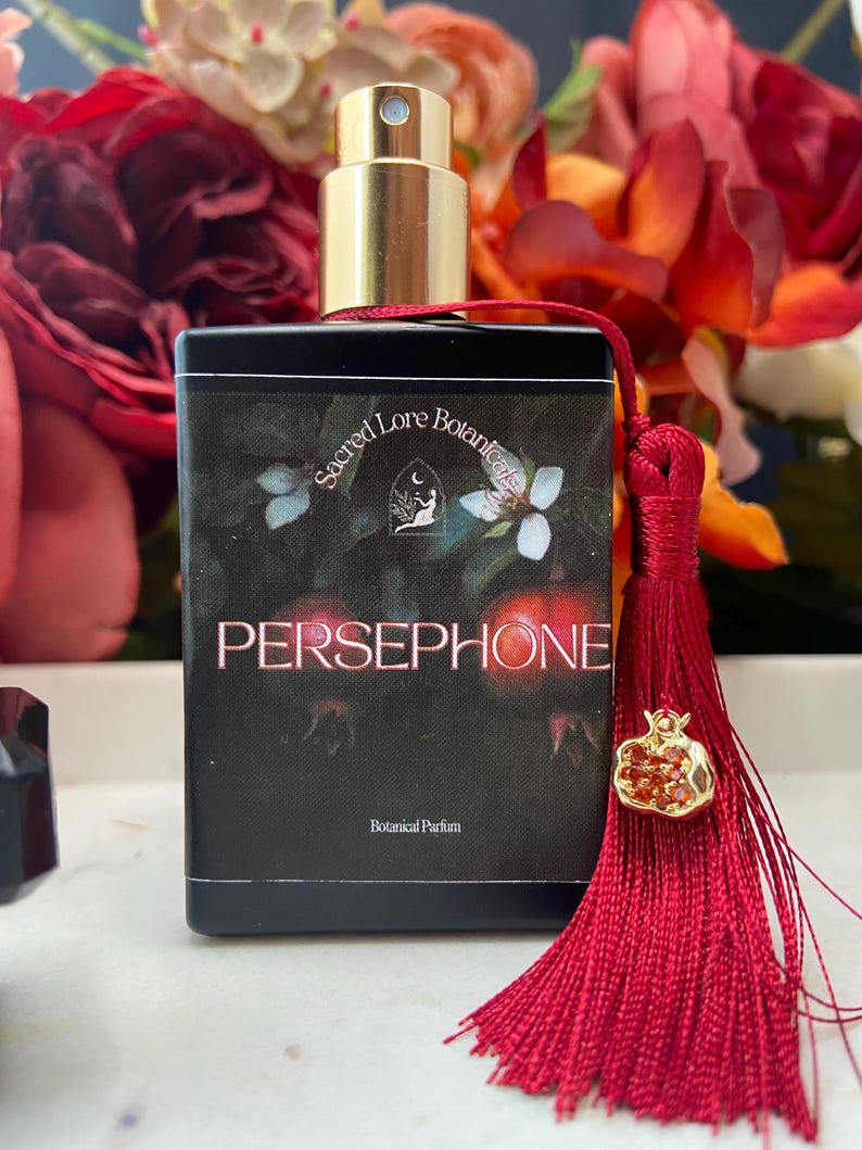 K&ouml;nnte beinhalten: Eine schwarze Parfumflasche mit goldener Kappe und rotem Quast. Die Flasche ist mit "Sacred Lore Botanicals Persephone Botanical Parfum" beschriftet.