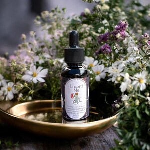 May include: A dark glass bottle with a black dropper, labeled "Uncord Me" with floral illustrations. The bottle sits on a gold-colored tray, surrounded by white and purple flowers. The product is a flower essence blend.
