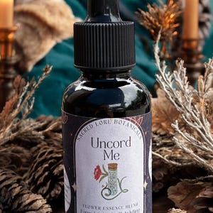 May include: A dark glass bottle with a black dropper and a label that reads "Uncord Me." The label features a floral illustration and text describing the product as a flower essence blend. The bottle is 2 oz / 60ml.