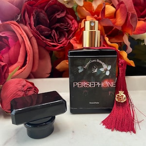 K&ouml;nnte beinhalten: Eine schwarze Glasflasche mit goldfarbenem Verschluss und rotem Quast. Die Flasche ist mit "Sacred Love Botanics" und "Persephone" sowie einer botanischen Illustration beschriftet. Die Flasche steht auf einer wei&szlig;en Oberfl&auml;che.