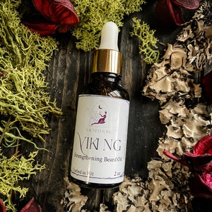 May include: A glass bottle with a gold cap and dropper top. The bottle is labeled "Sacred Lore Viking Strengthening Beard Oil, Crafted in NH, 2 oz."