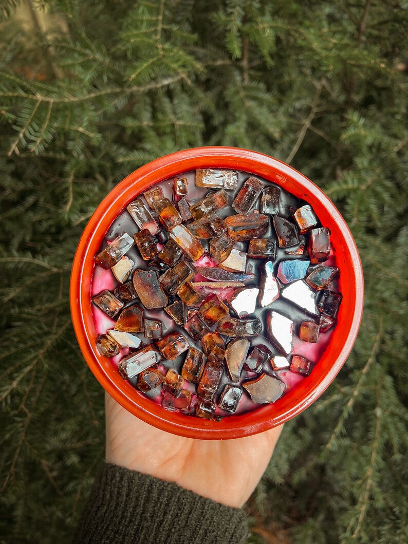 May include: A red candle jar with a black wax base and a layer of brown and silver glitter on top.