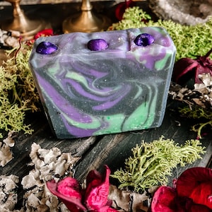 May include: A bar of handmade soap with a swirling pattern of green, purple, and black. The soap has three purple embellishments on top.