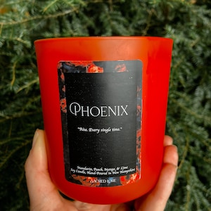 May include: A red soy candle with a black label that reads "PHOENIX" and "Rise. Every single time." The label also includes the text "Mandarin, Peach, Mango, & Lime Soy Candle, Hand-Poured in New Hampshire" and "SACRED LORE".