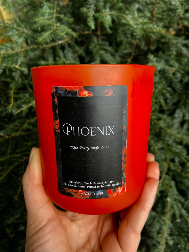 May include: A red soy candle with a black label that reads "PHOENIX" and "Rise. Every single time." The label also includes the text "Mandarin, Peach, Mango, & Lime Soy Candle, Hand-Poured in New Hampshire" and "SACRED LORE".