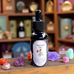 May include: A dark blue glass bottle with a black dropper, labeled "Uncord Me" with floral illustrations. The bottle is surrounded by colorful crystals on a wooden surface. The bottle contains 2 oz of flower essence blend.