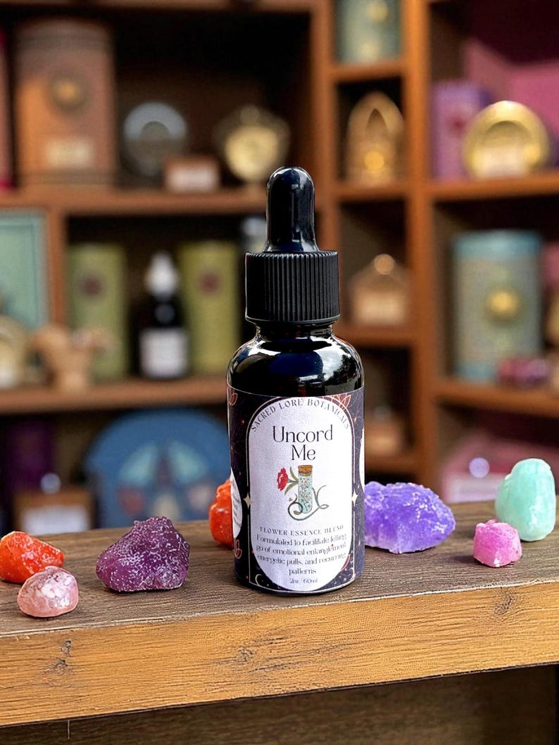May include: A dark blue glass bottle with a black dropper, labeled "Uncord Me" with floral illustrations. The bottle is surrounded by colorful crystals on a wooden surface. The bottle contains 2 oz of flower essence blend.