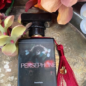 K&ouml;nnte beinhalten: Eine schwarze Glasflasche mit Parf&uuml;m, einem roten Quast und einem goldenen Apfel-Anh&auml;nger. Die Flasche ist mit "Sacred Love Botanicals Persephone Botanical Parfum" beschriftet.