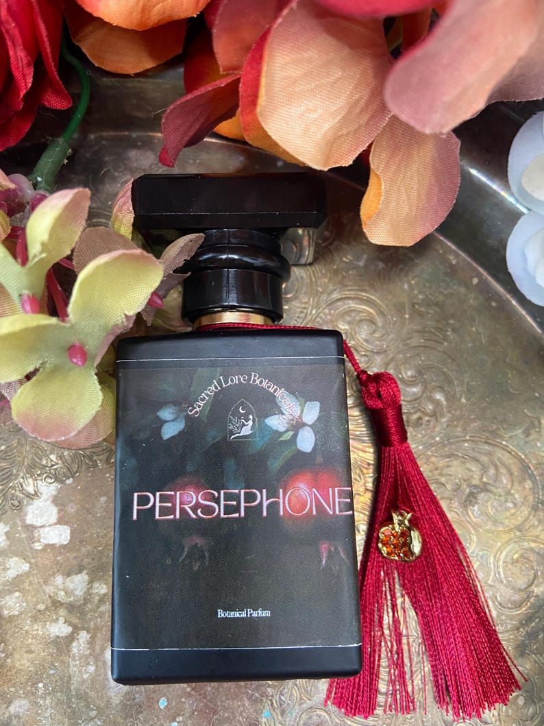 K&ouml;nnte beinhalten: Eine schwarze Glasflasche mit Parf&uuml;m, einem roten Quast und einem goldenen Apfel-Anh&auml;nger. Die Flasche ist mit "Sacred Love Botanicals Persephone Botanical Parfum" beschriftet.