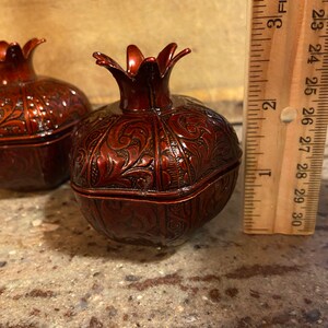 Puede incluir: Dos cajas de metal rojo en forma de granada adornadas con intrincados dise&ntilde;os florales. Las cajas miden aproximadamente 7,5 cm de alto y est&aacute;n sobre una superficie marr&oacute;n claro.