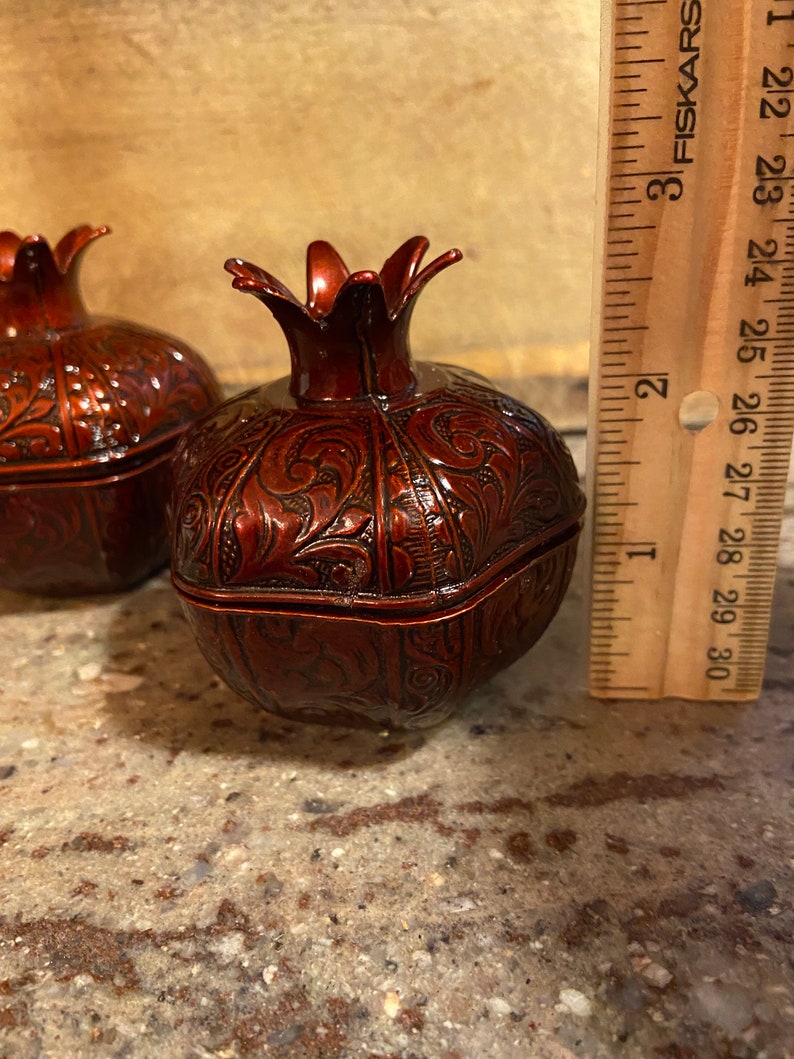 Puede incluir: Dos cajas de metal rojo en forma de granada adornadas con intrincados dise&ntilde;os florales. Las cajas miden aproximadamente 7,5 cm de alto y est&aacute;n sobre una superficie marr&oacute;n claro.