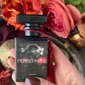 K&ouml;nnte beinhalten: Eine kleine schwarze Flasche mit einem roten Quast, beschriftet mit "Sacred Lore Botanics" und "PERSEPHONE" mit dem Text "Botanical Parfum".
