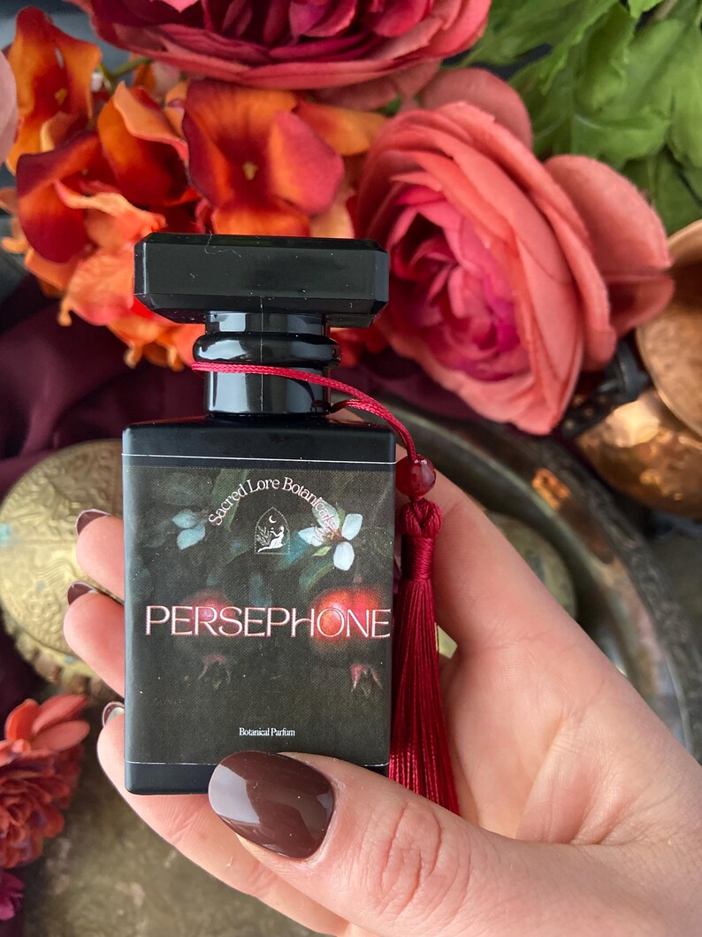 K&ouml;nnte beinhalten: Eine kleine schwarze Flasche mit einem roten Quast, beschriftet mit "Sacred Lore Botanics" und "PERSEPHONE" mit dem Text "Botanical Parfum".