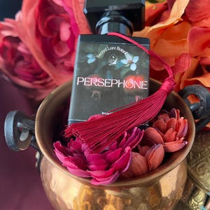 K&ouml;nnte beinhalten: Eine kleine Parf&uuml;mflasche mit einem roten Quast. Die Flasche ist schwarz mit einem goldenen Etikett, auf dem "Secret Lore Botanicals" und "PERSEPHONE" steht. Die Flasche ist von roten Blumen umgeben.