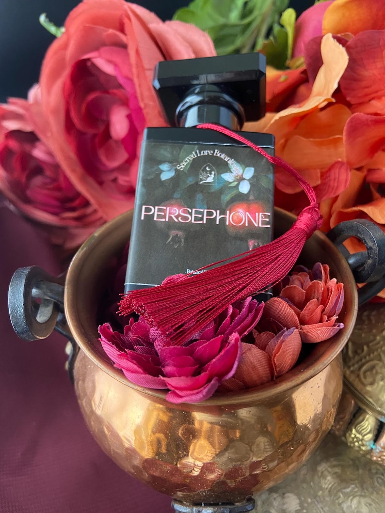 K&ouml;nnte beinhalten: Eine kleine Parf&uuml;mflasche mit einem roten Quast. Die Flasche ist schwarz mit einem goldenen Etikett, auf dem "Secret Lore Botanicals" und "PERSEPHONE" steht. Die Flasche ist von roten Blumen umgeben.