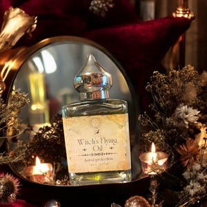 May include: A square glass bottle with a silver cap, labeled "Witch's Flying Oil - Astral projection." The bottle is surrounded by dried flowers, candles, and a mirror, set against a backdrop of deep red velvet. The label has a vintage aesthetic.