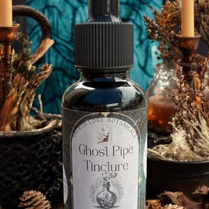 May include: A black glass bottle of "Ghost Pipe Tincture" with a dropper, featuring a label with botanical illustrations. The label reads "Sacred Lore Botanicals" and "Alcohol Tincture Crafted in New Hampshire". The bottle is surrounded by candles and dried plants.