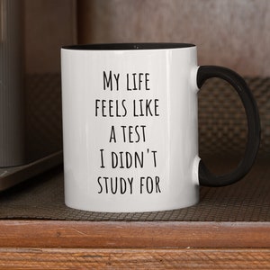 Puede incluir: Taza de café blanca con asa y borde negros. La taza tiene el texto "My life feels like a test I didn't study for" impreso en negro.