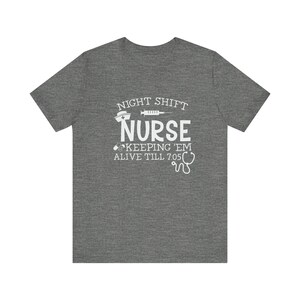 May include: A heather grey t-shirt with white text that reads "Night Shift Nurse Keeping Em Alive Till 7:05". The text is accompanied by a small graphic of a nurse's cap, a syringe, and a stethoscope.