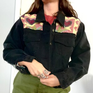 May include: A black denim jacket with a cropped fit and a colorful embroidered design on the back. The embroidery features hearts and geometric patterns in shades of red, yellow, green, and blue.