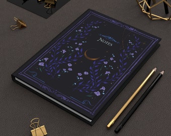 Può includere: Un quaderno nero con un design floreale e la parola "NOTES" in lettere dorate. Il quaderno ha una luna crescente e stelle sulla copertina. Il quaderno è su una superficie grigio scuro con due matite e graffette dorate.