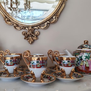 May include: A vintage tea set with gold accents, featuring a teapot, sugar bowl, and several teacups and saucers. The set has a white base with colorful, detailed artwork. A decorative gold mirror hangs in the background.