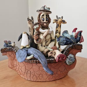 May include: A ceramic sculpture depicting Noah's Ark, with Noah, a giraffe, a penguin, an elephant, a cow, and other animals. The ark is reddish-brown with a textured surface. The animals are painted in various colours.