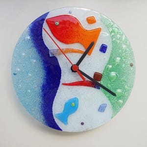 May include: A round, decorative wall clock with a colorful, abstract design. The clock face features a white background with blue, green, and red accents, and two fish shapes. The clock hands are black, with a red second hand.