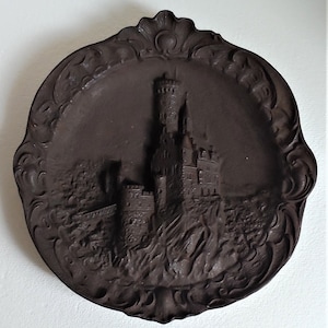 May include: A dark brown cast iron wall plaque featuring a 3D relief of a medieval castle with a tall tower. The plaque has a decorative border with a floral design.