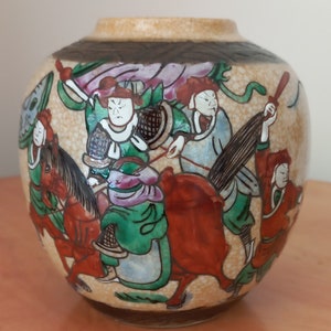 May include: A ceramic vase with a beige background and a brown rim. The vase is decorated with a colorful scene of people riding horses. The scene includes a pink, green, and blue flag.