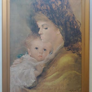 May include: An oil painting in a wooden frame shows a mother and child. The mother wears a black lace shawl and a yellow dress, holding the baby. The baby is wrapped in white fabric. The painting has a warm colour palette and is signed in the bottom right corner.