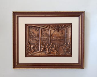 May include: A framed copper relief sculpture depicting a scene of people gathered in a room with a view of a cityscape. The sculpture is set against a white background.