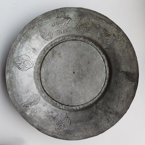 May include: A round, antique metal plate with a central recessed area. The plate features etched leaf and floral designs around the rim, along with text in an unknown language. The plate has a weathered, aged appearance.