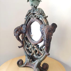 May include: A decorative mirror with an oval frame featuring two elephant figures on either side. The frame is made of a dark brown material with intricate carvings of leaves and vines. The mirror is topped with a small palm tree sculpture.
