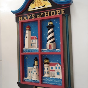 May include: A wooden wall hanging with a black frame and a red and blue interior. The interior features four windows with painted images of lighthouses and the text "RAYS of HOPE".