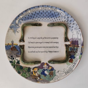May include: A round ceramic plate featuring four raised, green, fish-shaped figures arranged in a rectangle. The plate is decorated with blue fleur-de-lis, a coastal scene, and a group of figures in traditional clothing, along with French text.