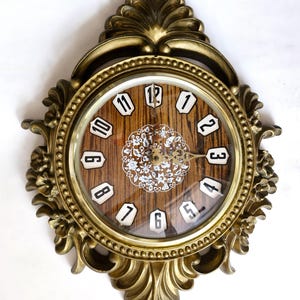 May include: Ornate gold-toned wall clock with a wood-grain face. The clock features black numerals, a decorative floral design, and a detailed, scrolling frame. The clock's face is approximately 15 cm in diameter.