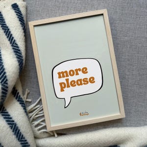 May include: A framed print with a light green background and the words "more please" in a speech bubble. The text is in a warm orange color. The frame is light wood. The print is resting on a gray surface next to a white and blue striped blanket.