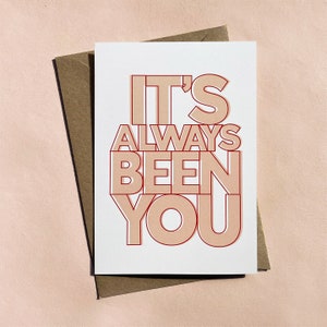 May include: A white card with the text "It's Always Been You" in red and peach colored lettering. The card is sitting on a brown envelope.