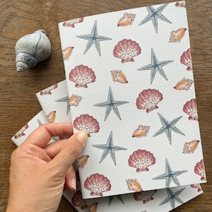 May include: A white notebook with a repeating pattern of seashells and starfish. The notebook has a white background with a light blue and brown pattern.