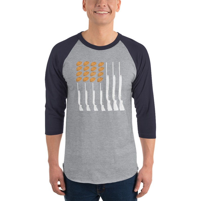 May include: A grey and navy blue baseball tee shirt with a white silhouette of an American flag. The flag is made up of shotguns and clay pigeons.