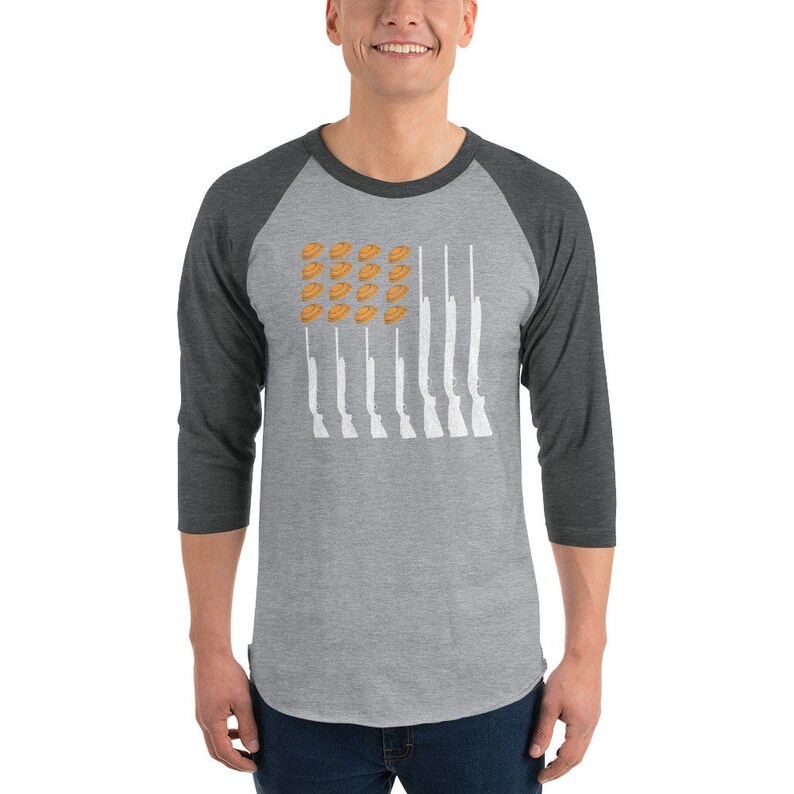 May include: A heather grey and charcoal grey baseball tee shirt with a white design of a flag with clay pigeons and shotguns.