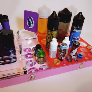 May include: A pink and clear organizer tray with various vaping supplies. The tray holds e-liquids, vape pens, and accessories. The tray has a skull and crossbones design on the front.